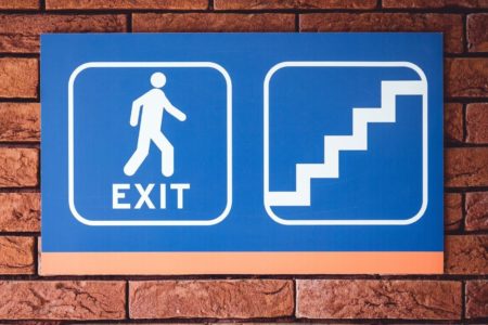 Et skilt montert på en mursteinvegg. Det blå skiltet viser Exit og en trappeoppgang.
I bunnen av skiltet er det en oransje strek som går langs hele skiltet